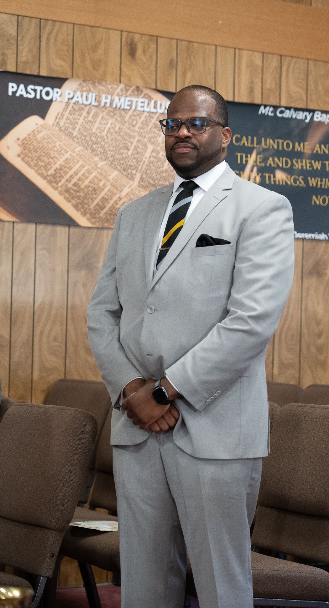 A person in a light gray suit and striped tie stands formally in a church setting with brown pews and a banner visible.