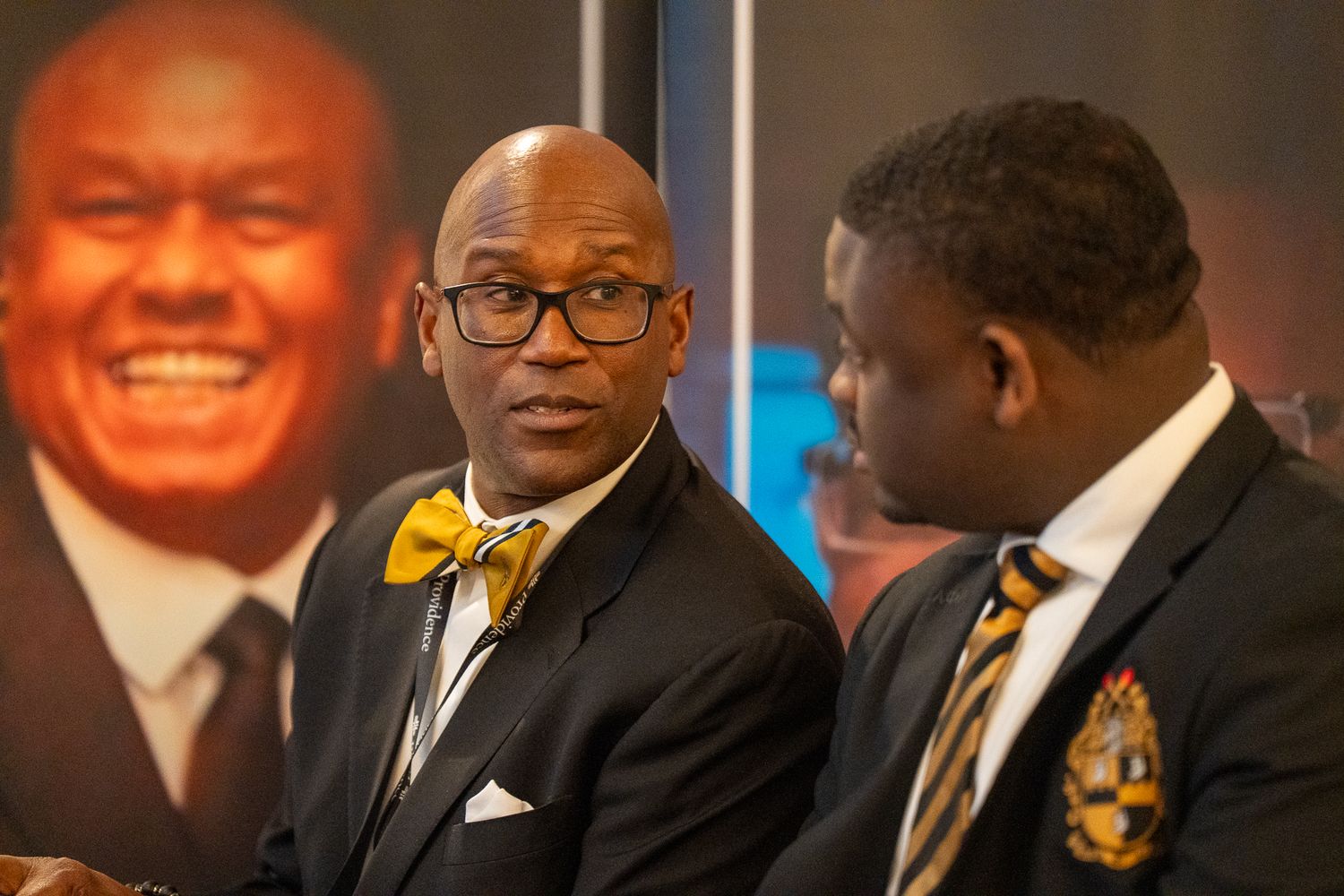 A formal event with multiple people wearing black suits and gold-accented ties seated at a table.