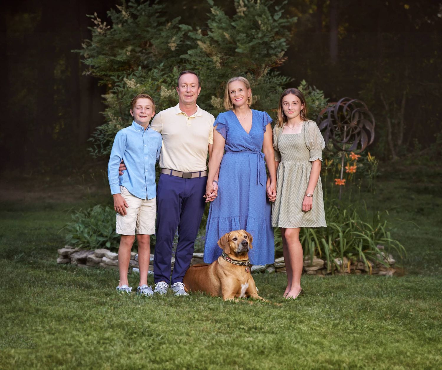 Parents and boy and girl tween holding hands and dog lying down in front of backyard planting at Joppa MD home