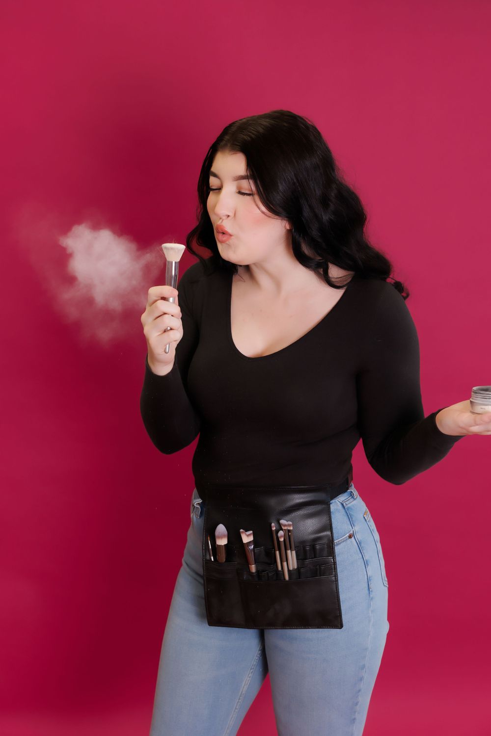 Makeup artist blowing powder with brush belt against pink background.
