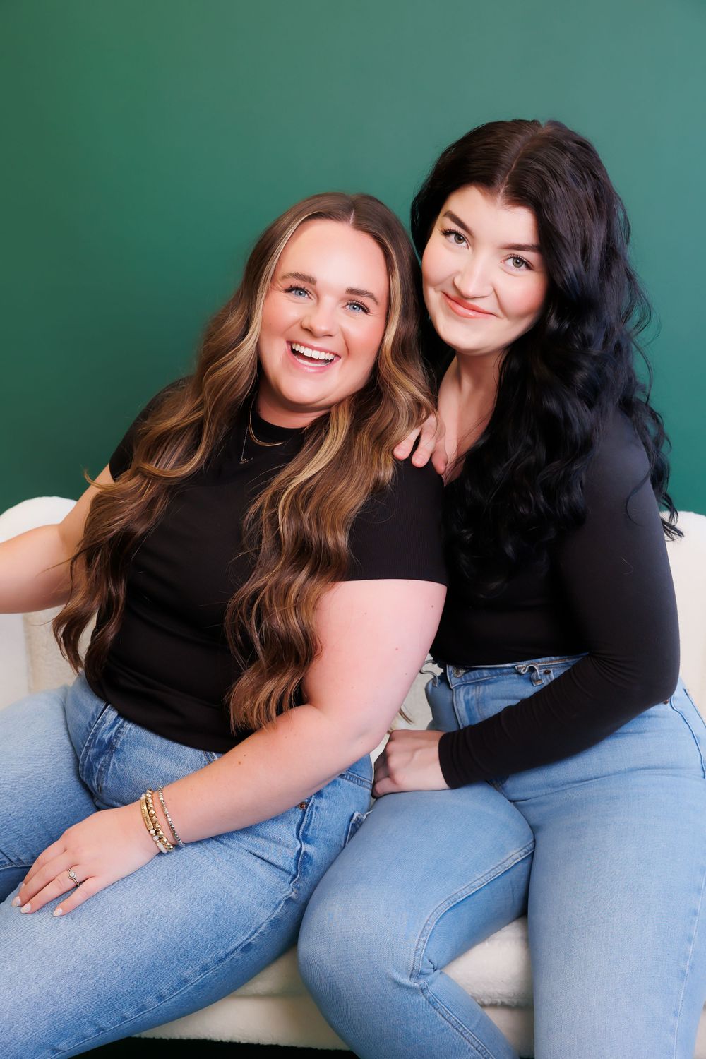 Two friends in matching black tops and jeans pose together against green background.