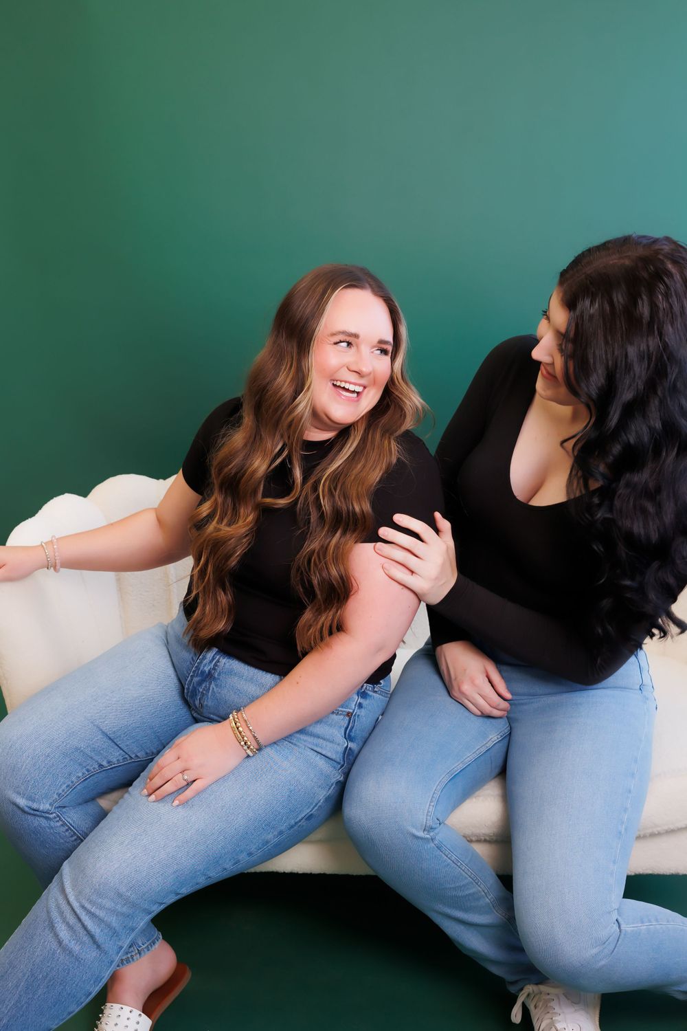 Two women sharing a joyful moment while sitting on white couch against green wall.