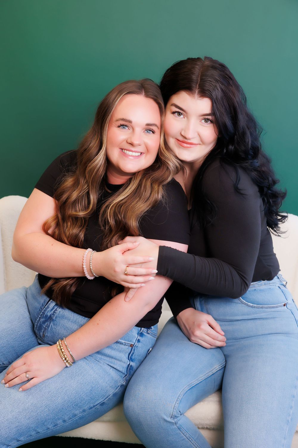 Two friends in casual outfits share a warm embrace on a white couch against a green background.