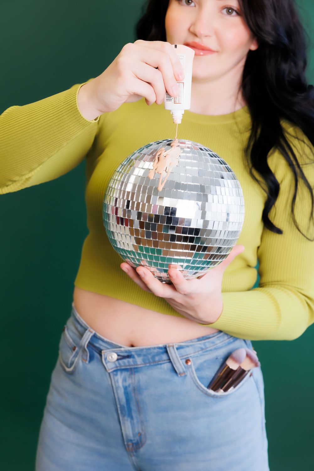 Person in yellow sweater applies glitter to disco ball against green backdrop.