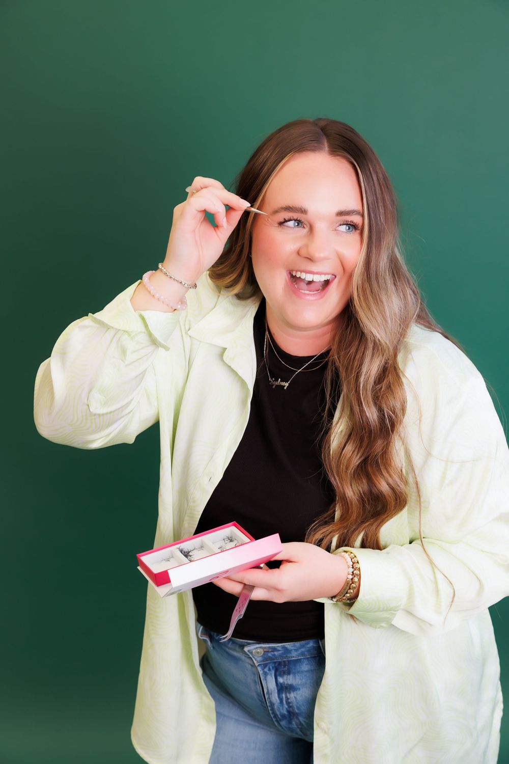 Person in light jacket applying makeup while smiling against green background.