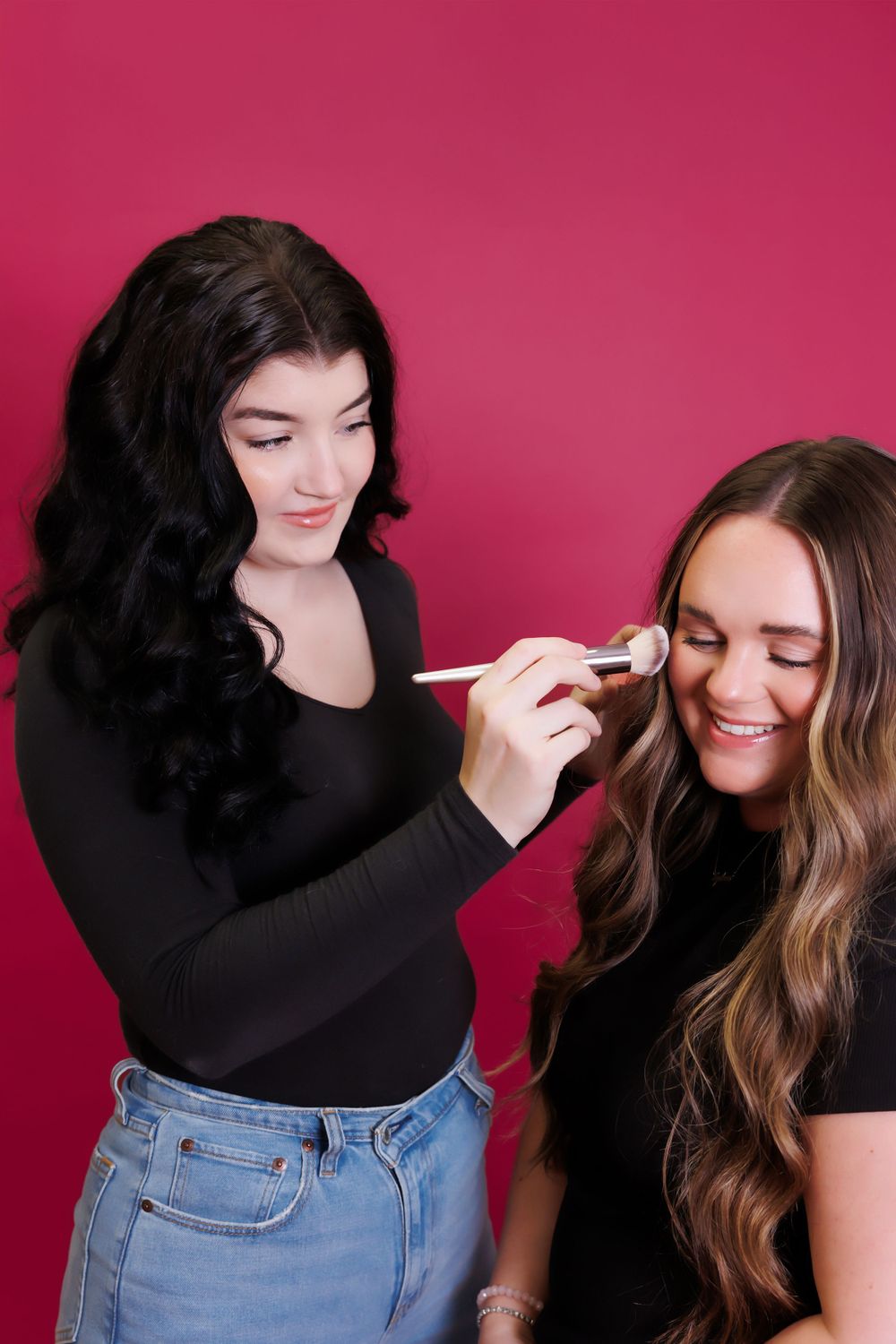 Two friends during makeup application session against vibrant pink background.
