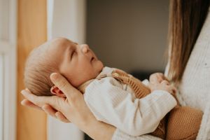 A sleeping infant wearing a cream-colored sweater is cradled gently in someone's hands.