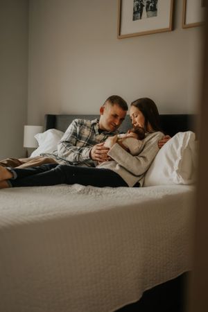 A couple cuddles with their newborn baby on a white bed.
