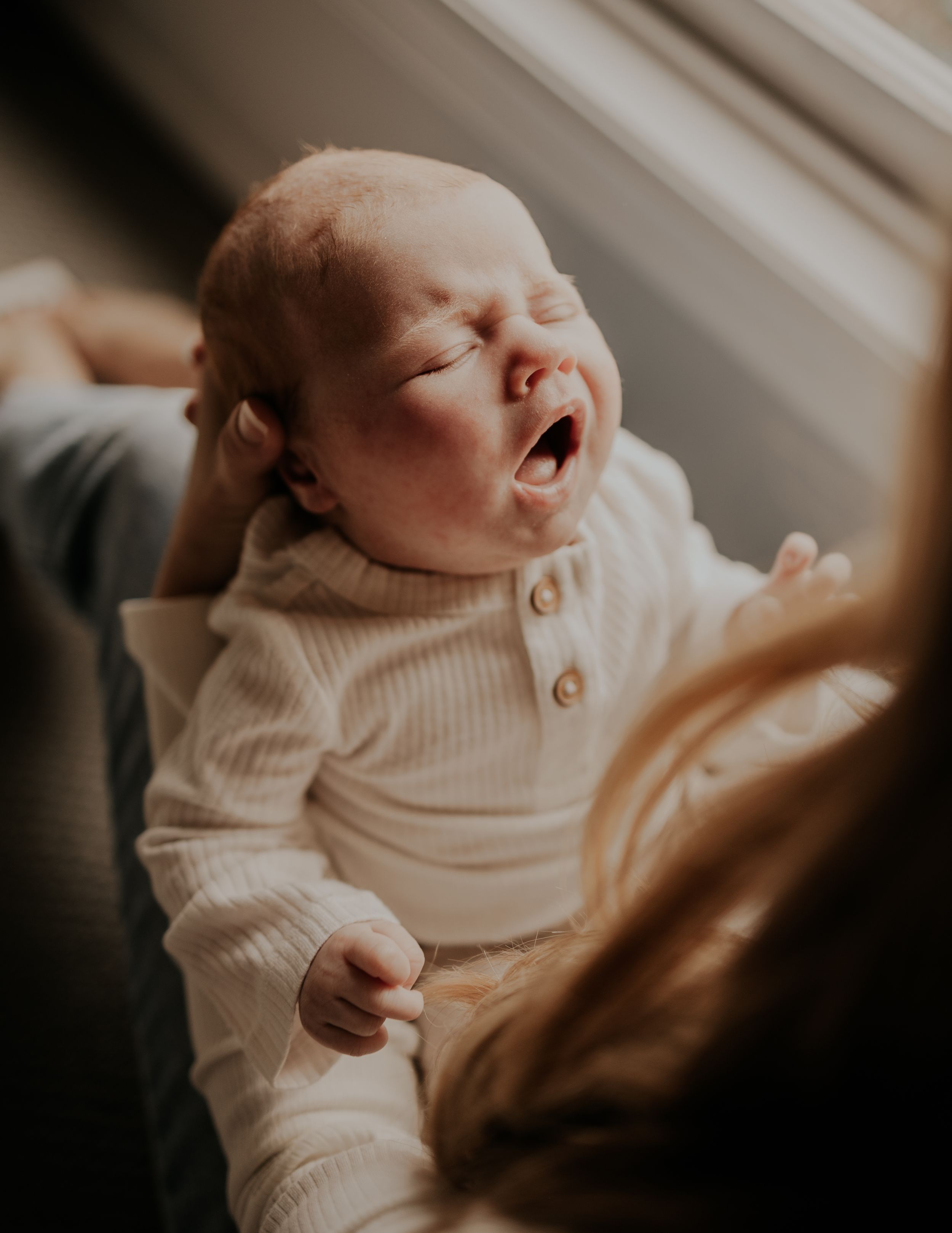 A sleepy newborn baby yawns while wearing a cream colored ribbed outfit.