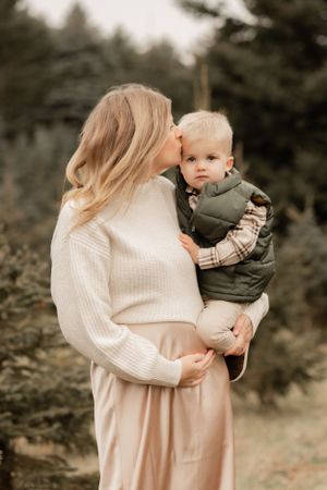 A pregnant woman in a white sweater kisses her toddler in a winter outdoor setting.