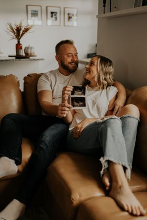A couple shares a joyful moment on a leather couch while holding an ultrasound photo.