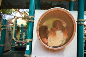 Two kids shown in blurred view through a circular porthole window on a playground structure.
