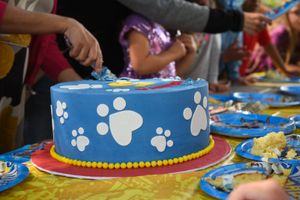 A blue birthday cake decorated with white paw prints sits on a colorful party table surrounded by blue plates.