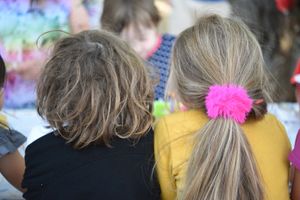 Two kids seated together at an event, with one wearing a bright pink hair tie in their ponytail and yellow shirt. Children's birthday photo.