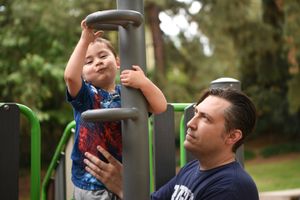 Child plays on outdoor fitness equipment while adult supervises in a park setting at a birthday party.
