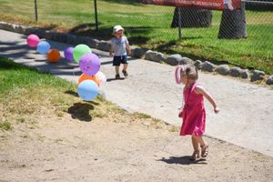 Children playing with colorful balloons on a gravel path in a sunny outdoor setting at a birthday party.