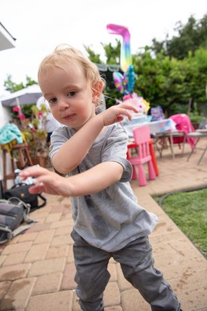 A small child in gray clothing plays outside on a patio with colorful party decorations visible in the background.