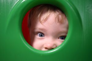Toddler peeks playfully through a round green opening in a playground structure on a birthday party.