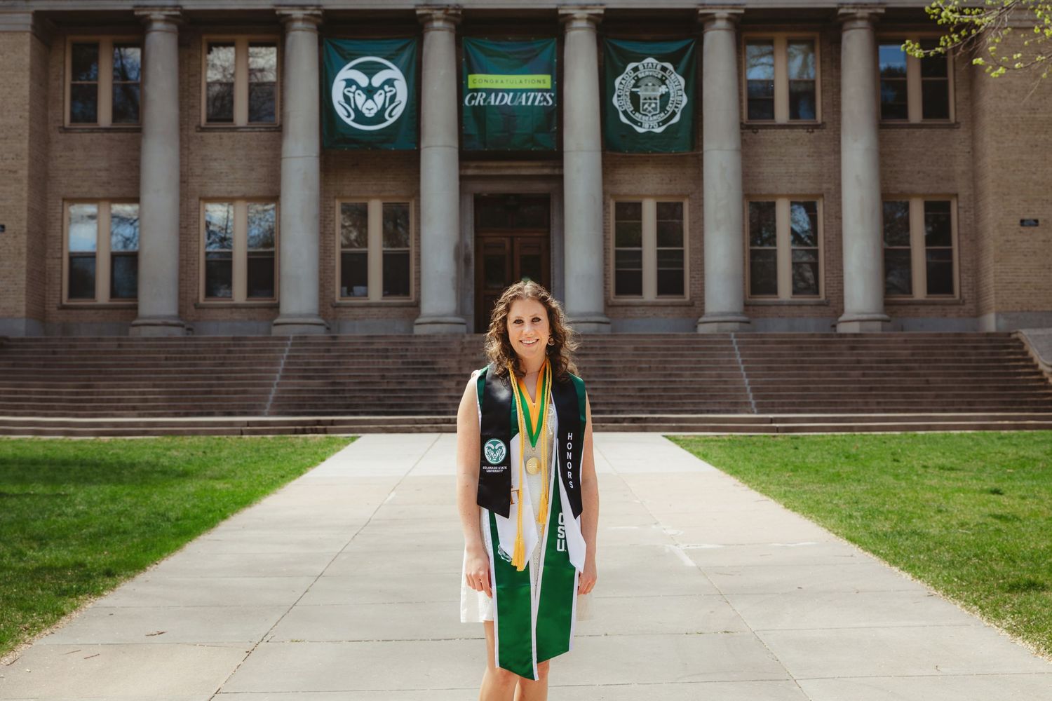 Graduate in green and white regalia standing in front of Colorado State University administration building.