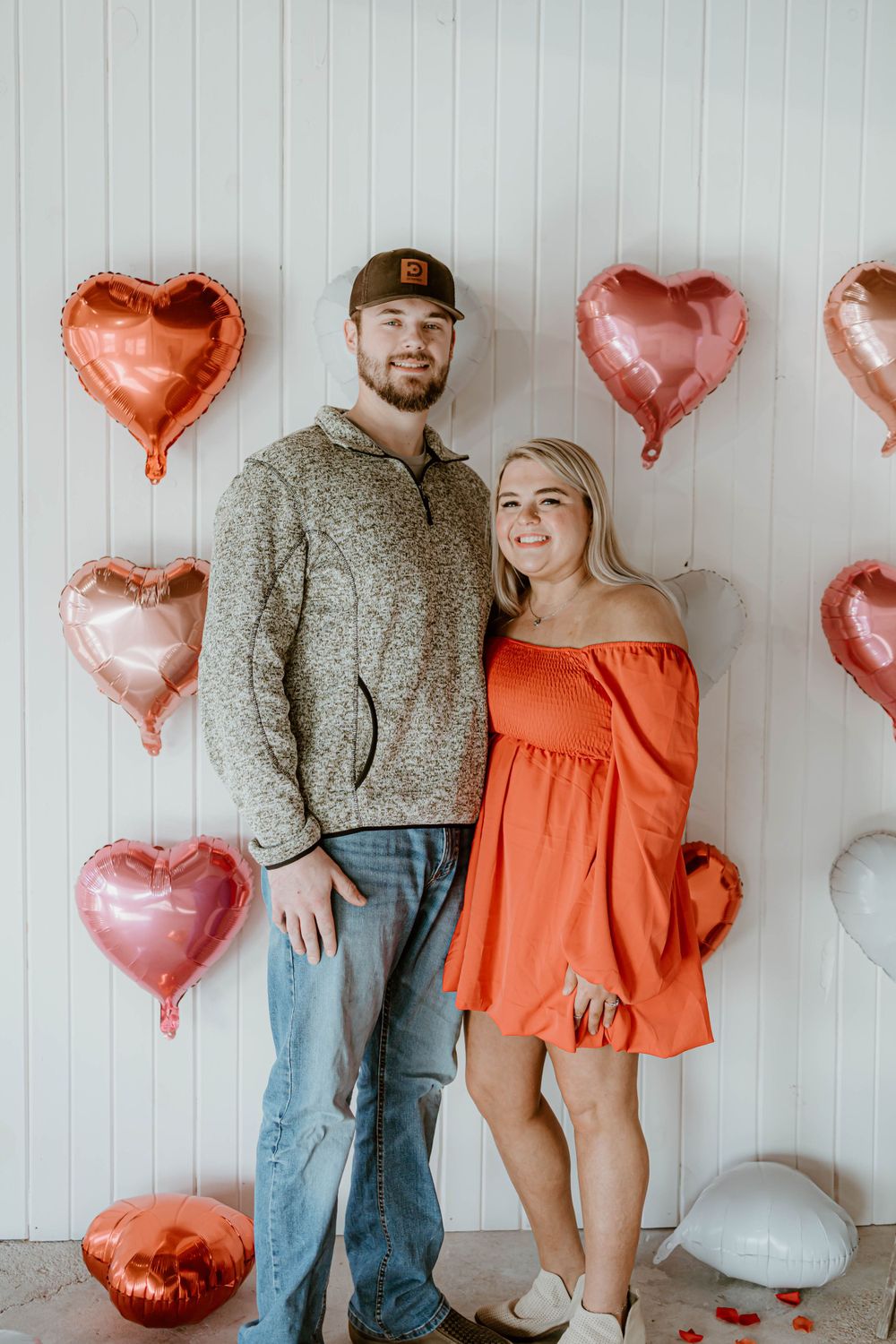 Couple posing in front of rose gold heart balloons with orange dress and grey sweater.
