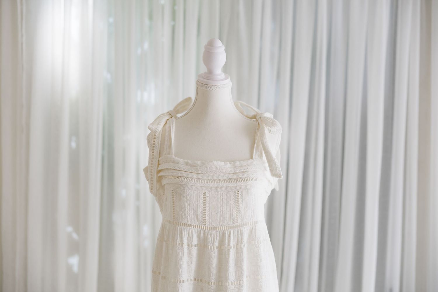 Vintage white cotton dress with delicate shoulder ties displayed on a mannequin against light curtains.