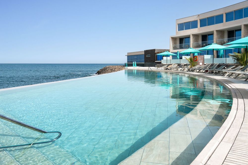 Luxury infinity pool overlooking the ocean with modern hotel rooms and lounge chairs along the curved edge.