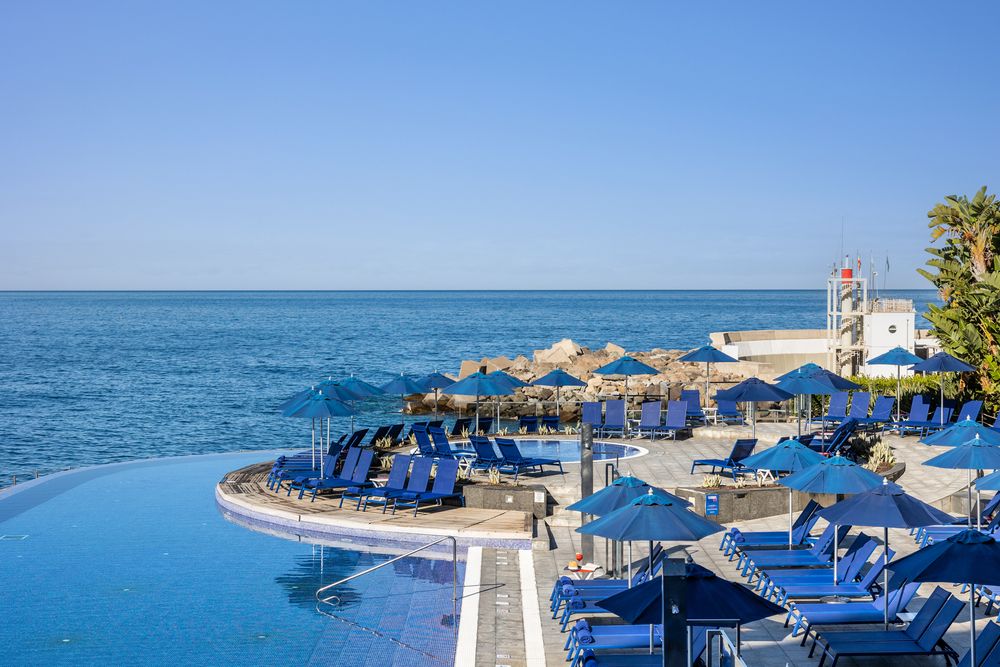 Luxury infinity pool with blue loungers and umbrellas overlooking the sparkling Mediterranean Sea on a clear sunny day.