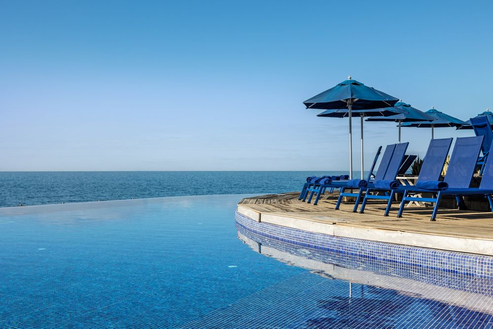 Luxury infinity pool with blue lounge chairs and umbrellas overlooking a calm ocean under a clear blue sky.