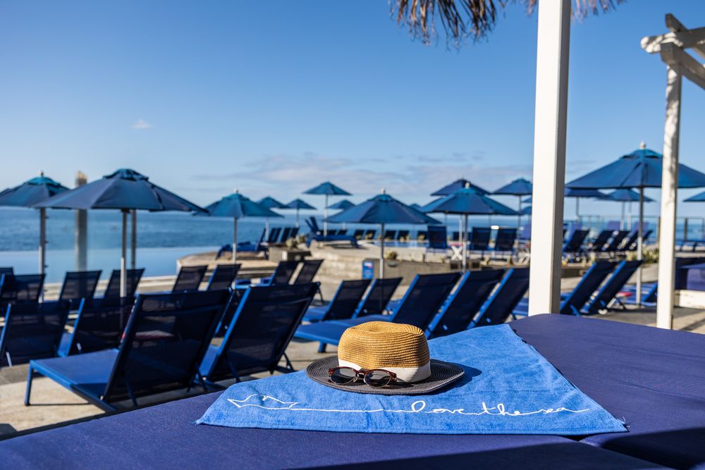 Blue lounge chairs and umbrellas on a sunny beachfront with a straw hat and sunglasses on a towel.