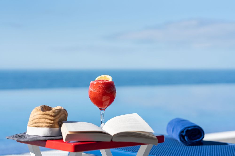 A refreshing red cocktail in a wine glass beside an open book, straw hat, and blue towel by an infinity pool.