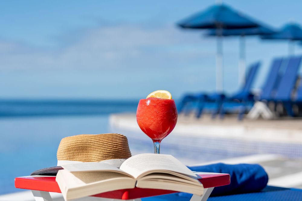 Tropical poolside scene with a red cocktail, open book, and straw hat by an infinity pool with blue umbrellas.