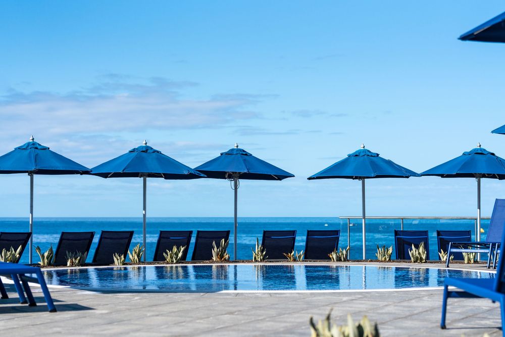 Luxury resort pool with blue lounge chairs and umbrellas overlooking a calm ocean under a clear blue sky.