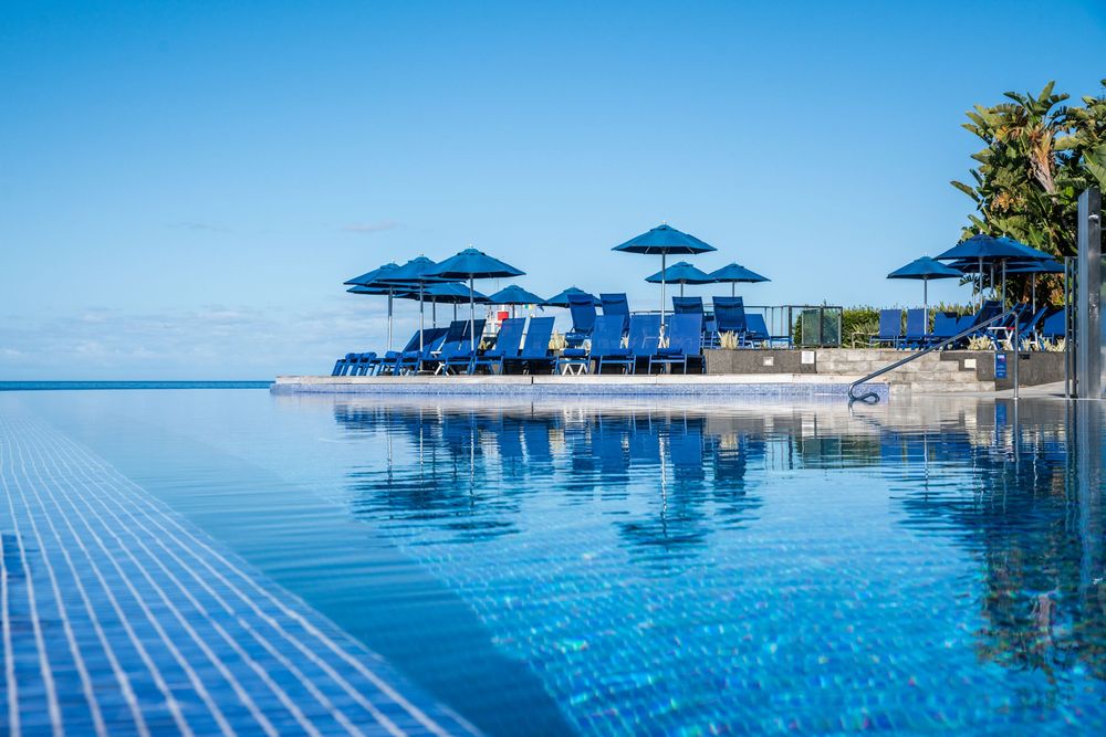 Luxury infinity pool with blue lounge chairs and umbrellas overlooking a calm ocean under a clear blue sky.