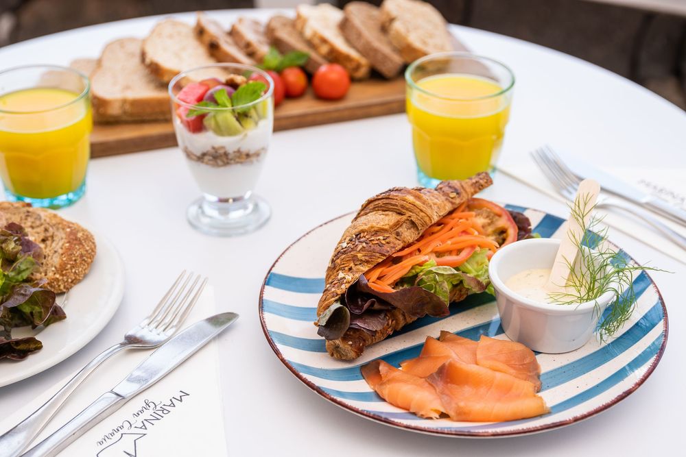 A gourmet breakfast spread with smoked salmon croissant, yogurt parfait, bread board, and fresh orange juice on a white table.
