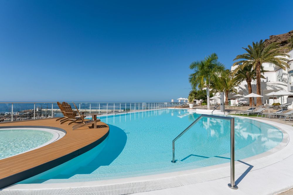 Luxurious outdoor infinity pool with turquoise water, wooden deck, palm trees, and stunning ocean views under a clear blue sky.