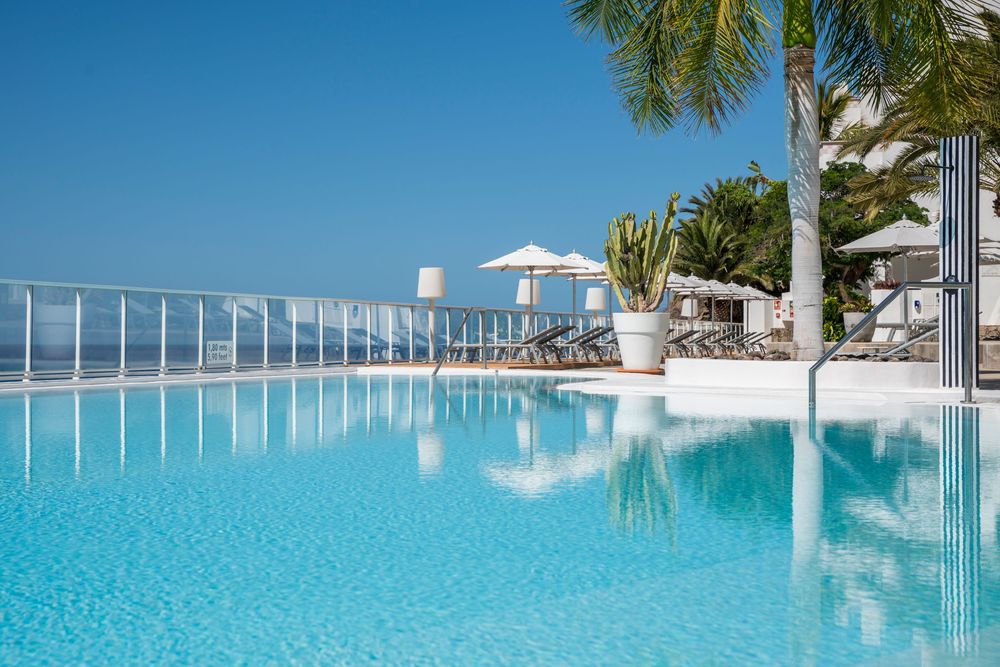 Luxury resort infinity pool with crystal-clear blue water, palm trees, and ocean views under a bright blue sky.