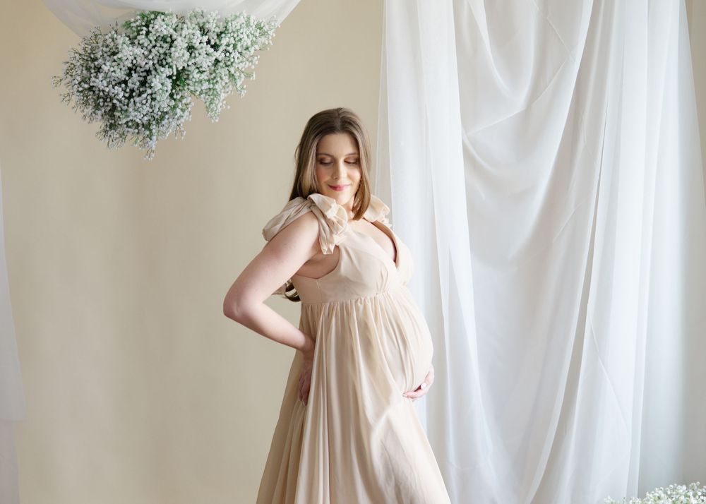 Studio-style maternity photo, woman in beige gown holding baby bump with floral backdrop.