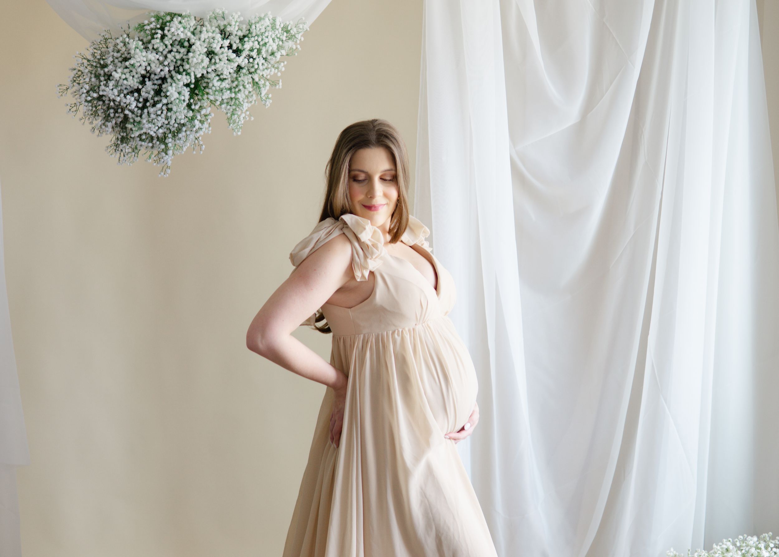Studio-style maternity photo, woman in beige gown holding baby bump with floral backdrop.