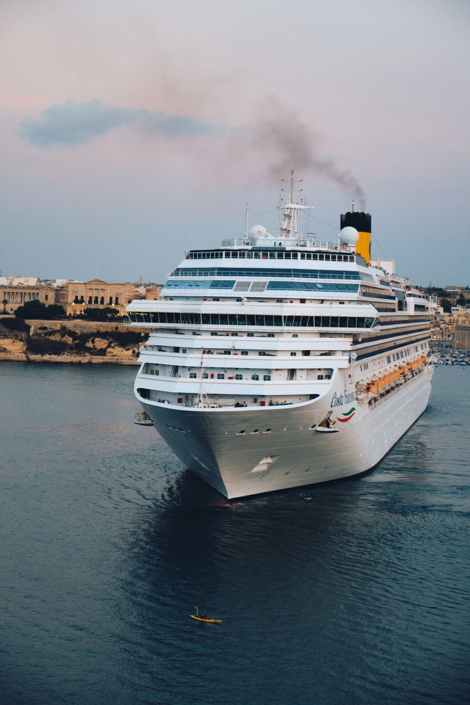 A white cruise ship with smoking funnel docked in a Mediterranean port.