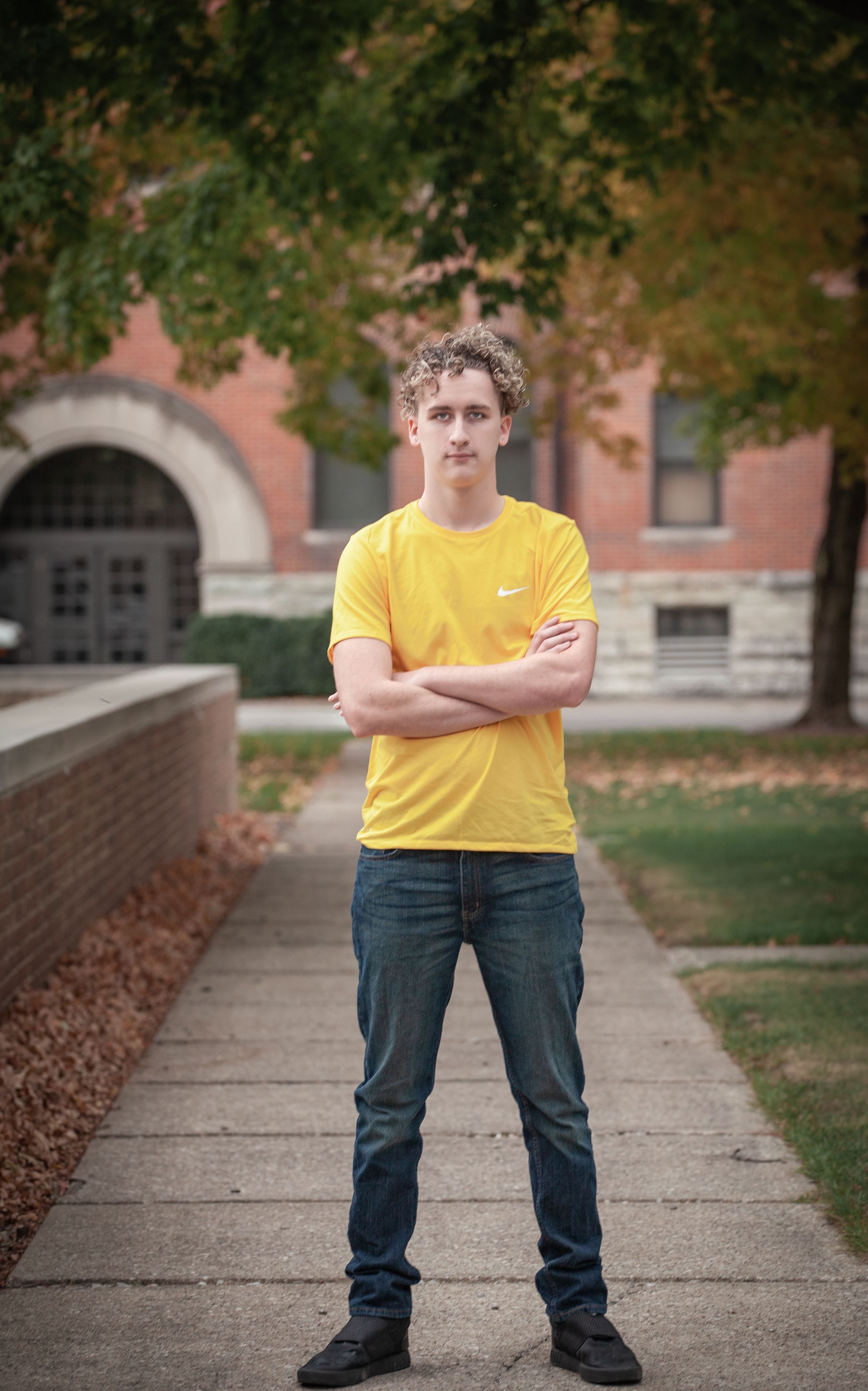Person in yellow t-shirt standing on sidewalk with brick building and fall trees behind.