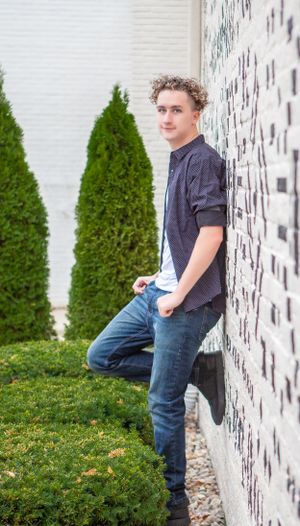Casual outdoor portrait against a white wall with evergreen shrubs in the background.