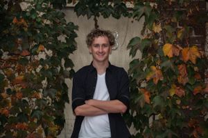 Person in a navy polka dot shirt poses against a wall with autumn foliage.