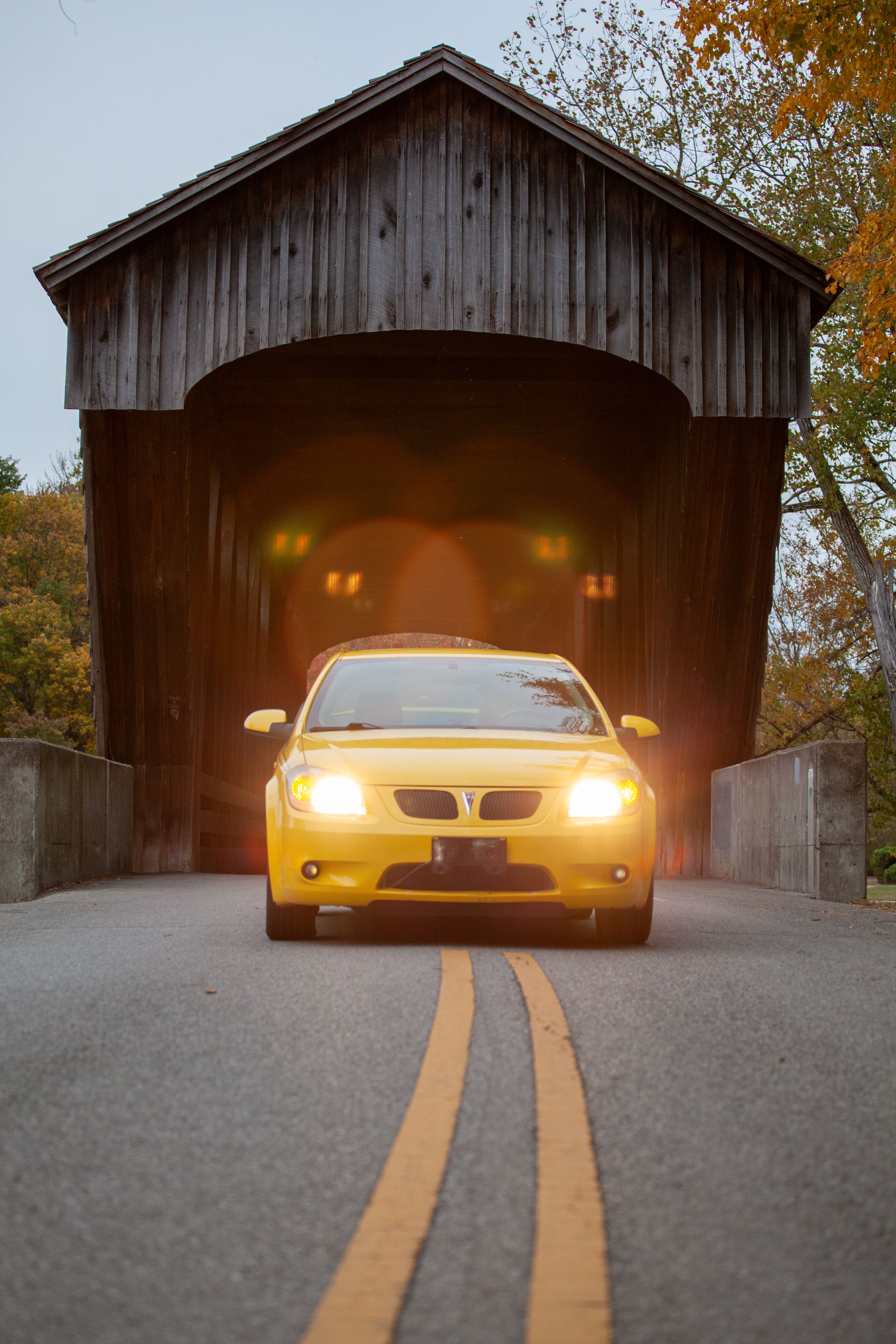 Yellow headlights shine through covered bridge at dusk with car silhouette.