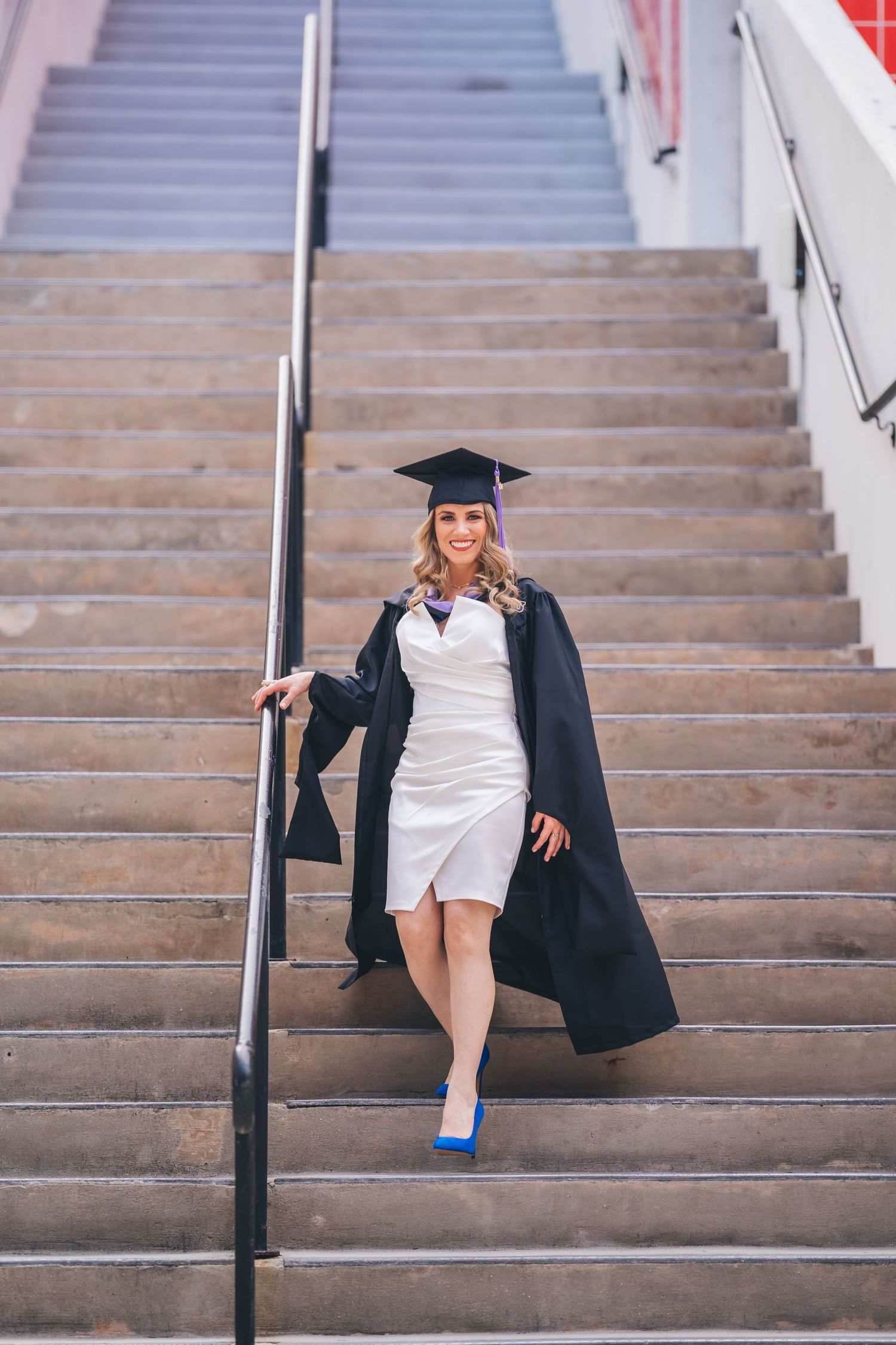 Graduate in cap and gown poses on concrete stairs wearing a white dress and blue heels.