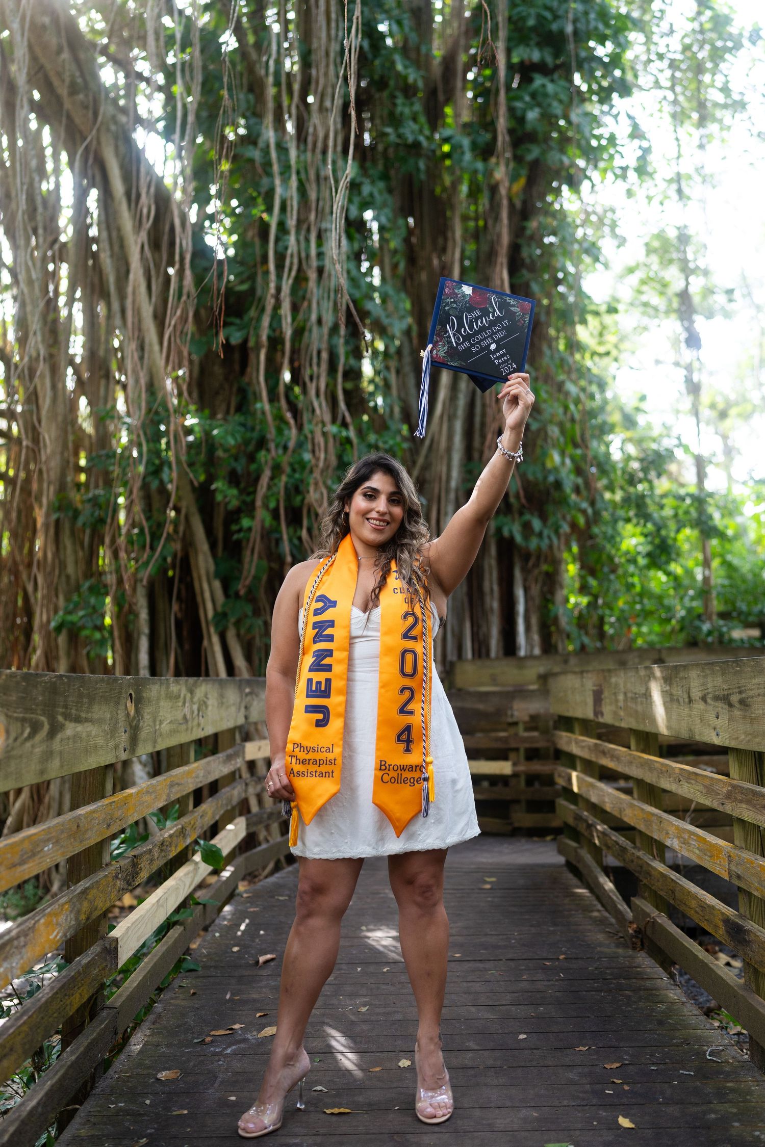Graduate in blue and gold stole celebrates on wooden boardwalk surrounded by banyan trees.