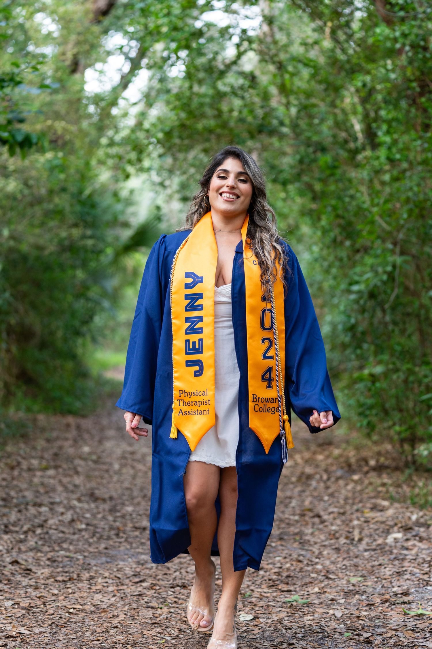 Graduate walking in blue cap and gown with gold graduation stole on nature path.