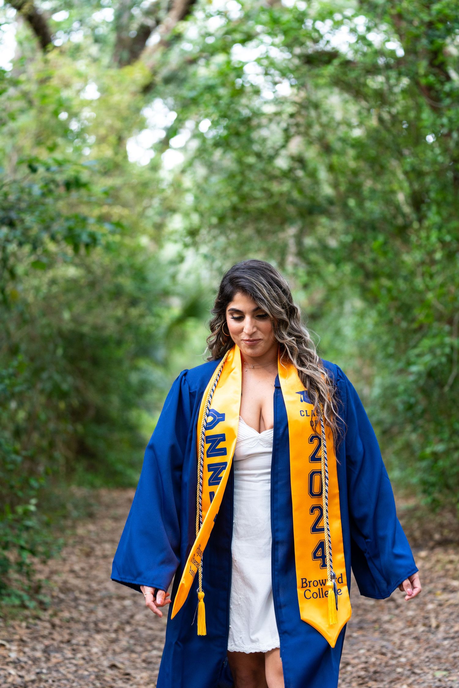Graduate in blue graduation gown and gold stole poses on forest trail.