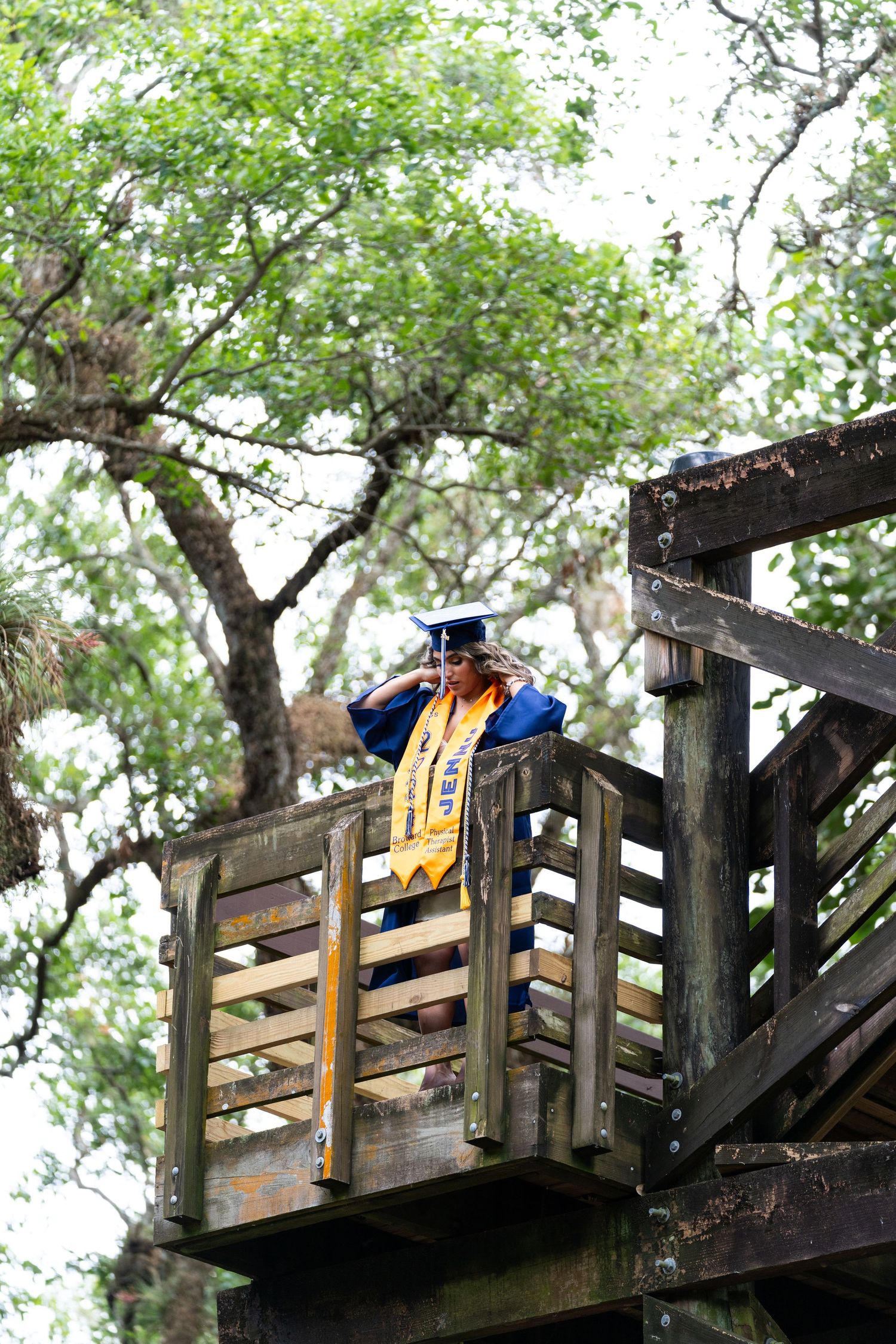Graduate in blue and gold regalia stands on wooden deck platform surrounded by trees.