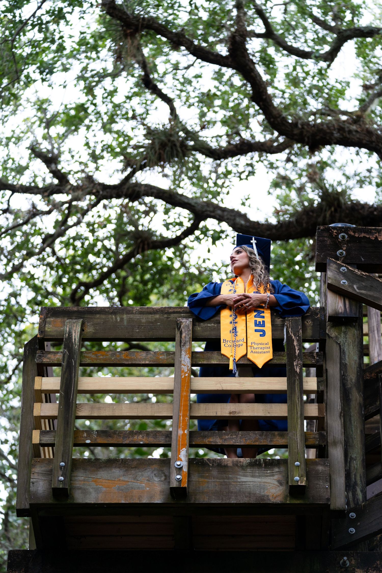 Graduate wearing academic regalia and stoles poses on rustic wooden platform with trees in background.