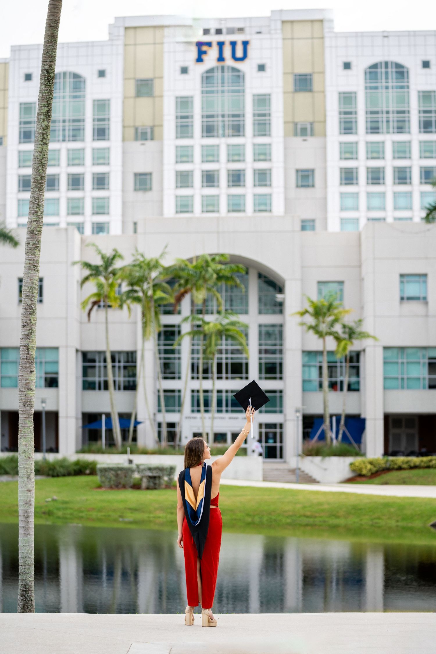 FIU graduate in red gown poses in front of campus building with reflection pool.